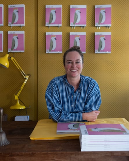 Annabelle Hickson, a woman wearing a checked blue and white shirt with brown hair, is sitting at a desk. In front of her, and also hanging behind her on a mustard-yellow wall, are copies of Galah magazine, pink with a white and grey illustration of a bird on the cover.
