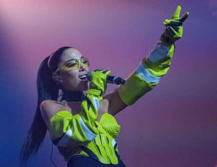 A woman with a long black ponytail and a fluorescent top and a conical bra. She is singing and holding up her right arm