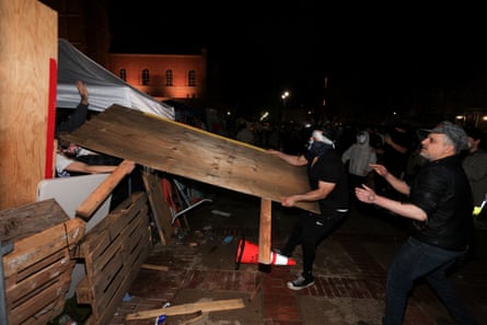 Person in black holds a wood plank as another person dressed in black stands in the back