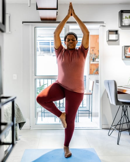 A relaxed-looking woman in a dusty red-T-shirt and maroon leggings holds the tree pose. Her arms are above her head, her hands in a prayer-like position; one leg is bent and resting on the standing leg