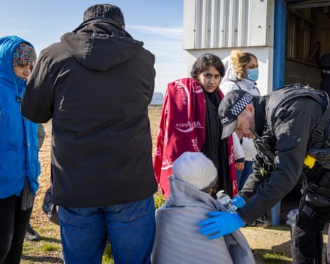 A police officer gives water to a young boy on a beach, with others of varying ages stood around them