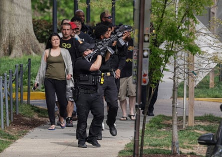 Police escort people down a tree-lined street with guns drawn and aimed at a point above and behind them.