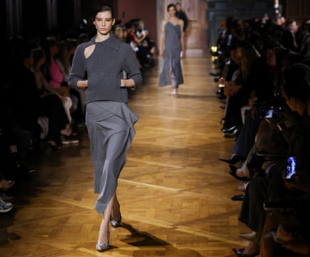 A model wearing a structured grey midi skirt and long grey top with cutouts around one shoulder.