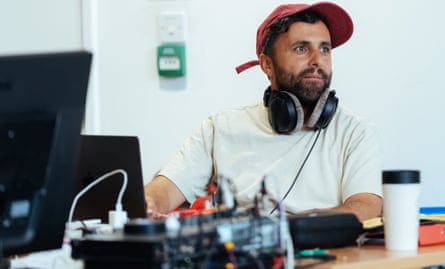 Composer Dom Coyote sits at a desk with headphones round his neck, near a laptop and cables