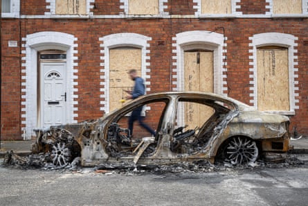 A burnt-out car in front of boarded up houses