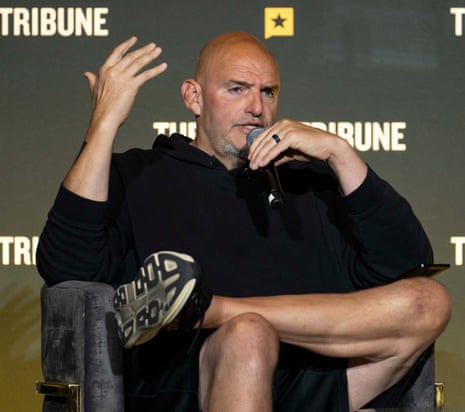 a man gestures while speaking into a microphone, seated with feet up