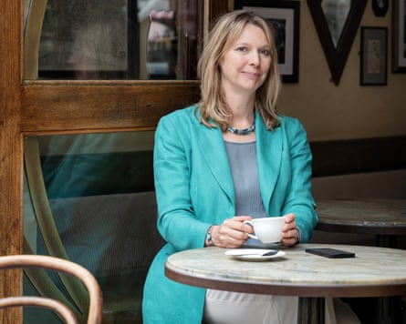 A woman in a teal jacket over a grey top sits at a circular table in a coffee shop