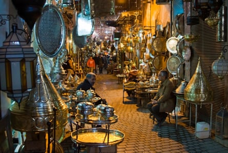 Two men sat outside in a Moroccan souk, surrounded by gold objects during the nighttime.
