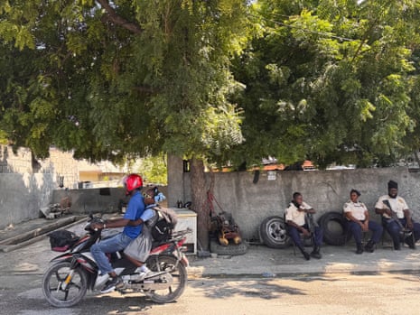 A line of armed, uniformed police officers sit on plastics chairs as a man and a child pass by on a scooter