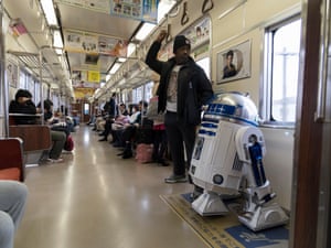 Richard, standing on a Japanese train and looking out the window, next to his R2-D2-like android