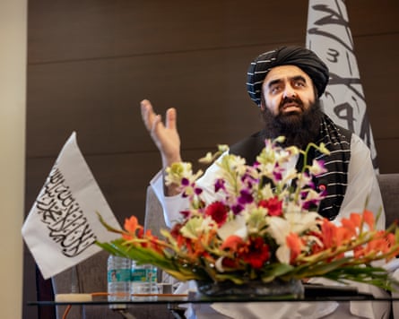 Afghanistan’s foreign minister Mawlawi Amir Khan Muttaqi wearing a black turban and traditional clothing speaks behind a table decorated with flowers, with Taliban flags visible in the background