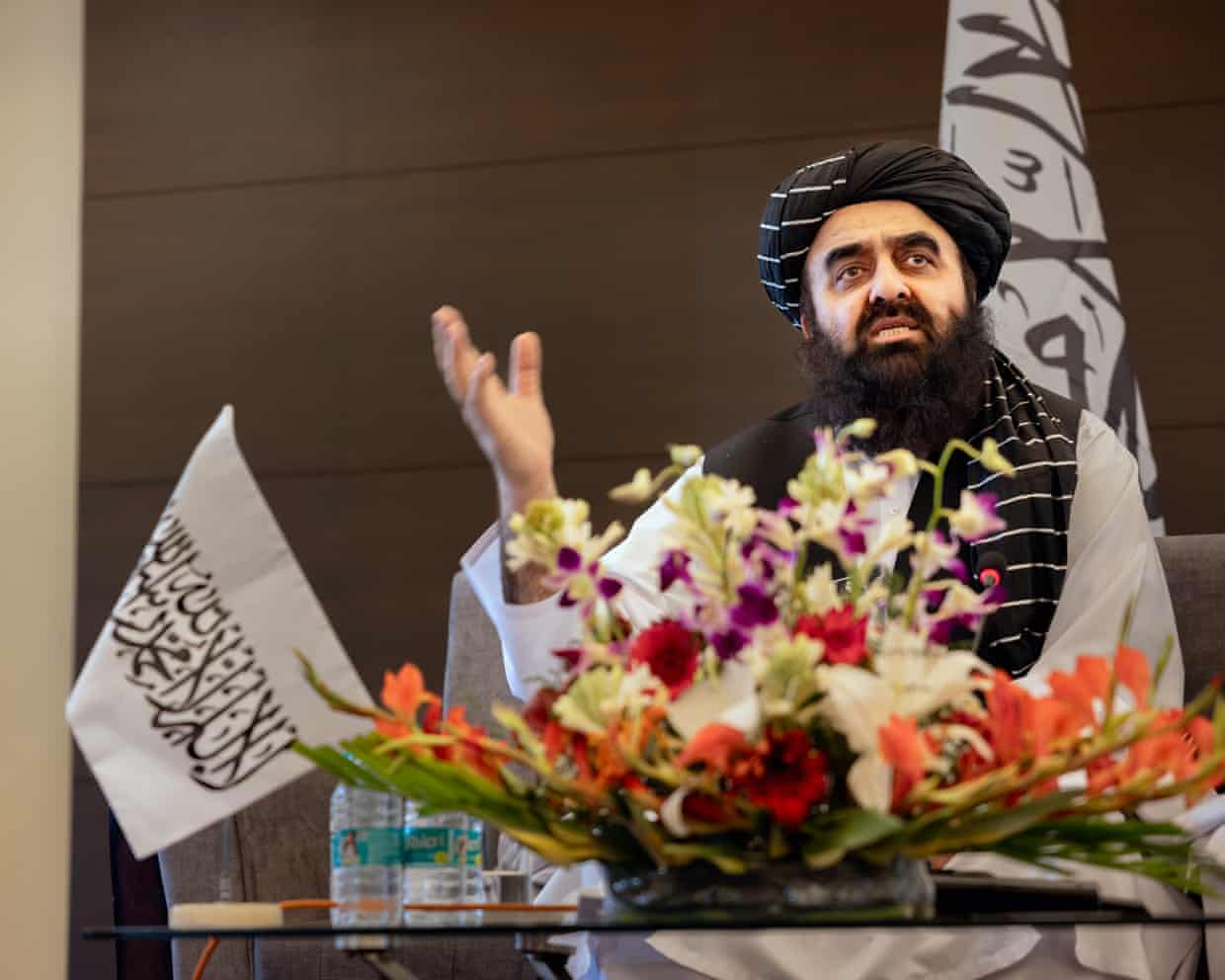Afghanistan’s foreign minister Mawlawi Amir Khan Muttaqi wearing a black turban and traditional clothing speaks behind a table decorated with flowers, with Taliban flags visible in the background