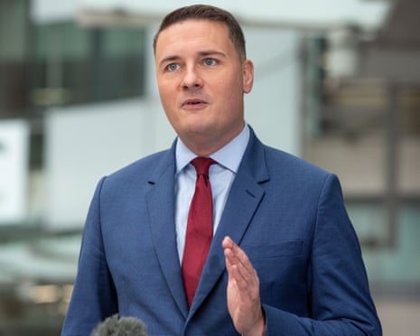 Wes Streeting in a blue suit and red tie speaks while gesturing, with a microphone visible below
