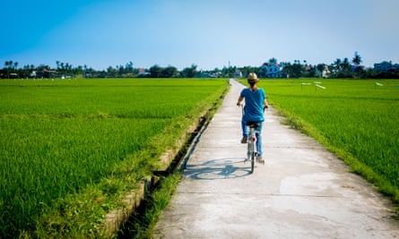 Rear view of a woman riding a bicycle on a footpath amidst farm, in Hoi An, Vietnam.