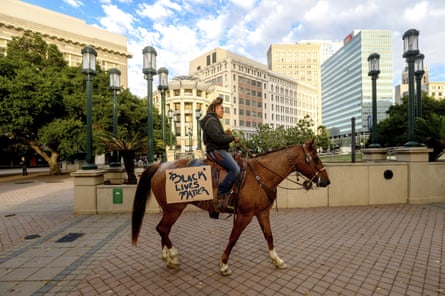 Young Black woman wearing jeans and a hooded Black sweatshirt rides brown horse with cardboard sign on flank that says Black Lives Matter across brick plaza, with high-rise buildings in the background.