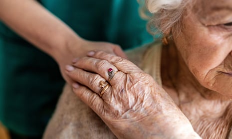 An elderly woman holds the hand of a care worker, which is resting on her shoulder