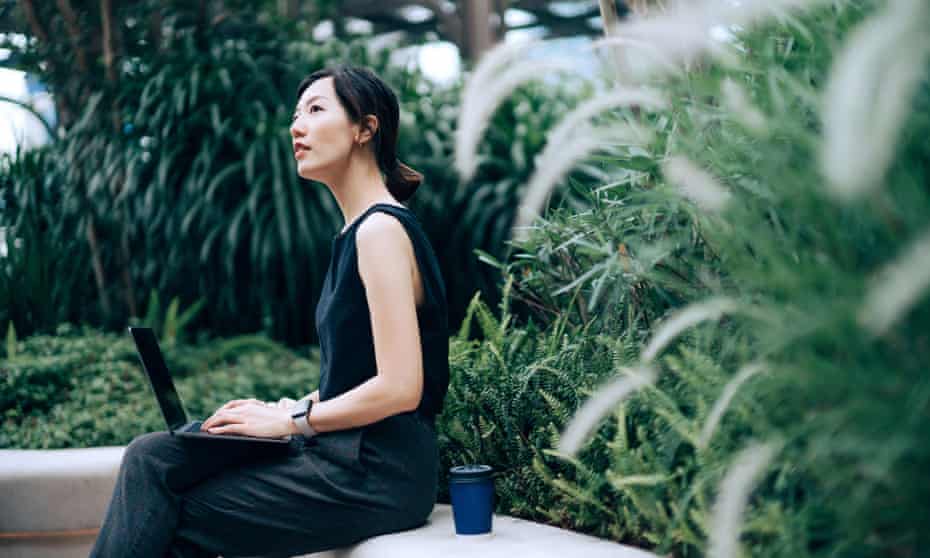 Young Asian businesswoman working on laptop outdoors, sitting on the bench in an urban office park, surrounded by green plants. 