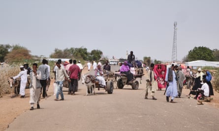 A stream of people, some walking, some on carts pulled by donkeys, move along a wide, dusty road.