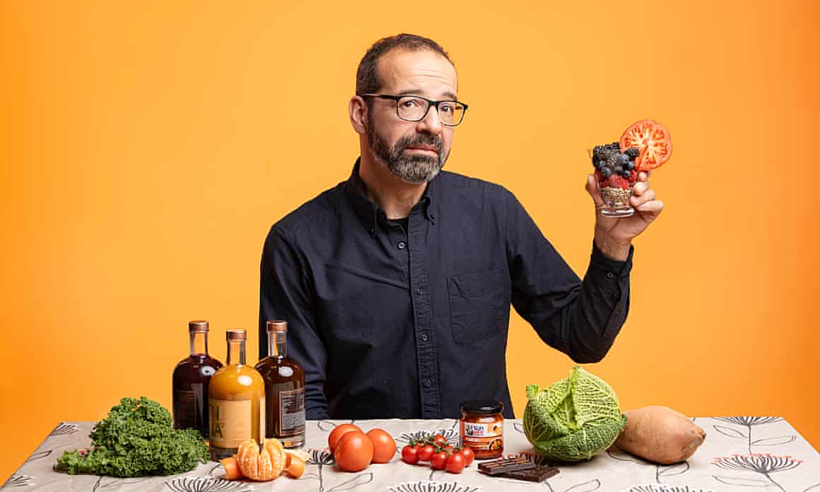 Steve Rose sits at a table filled with fruit and veg holding up a glass