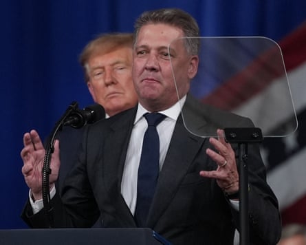 Donald Trump watches from behind as Gernot Döllner speaks at a lectern