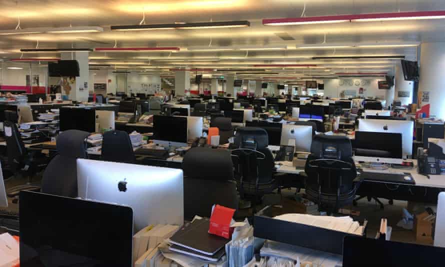 Photograph looking across rows of empty desks with computers under strip lights in the Guardian newsroom