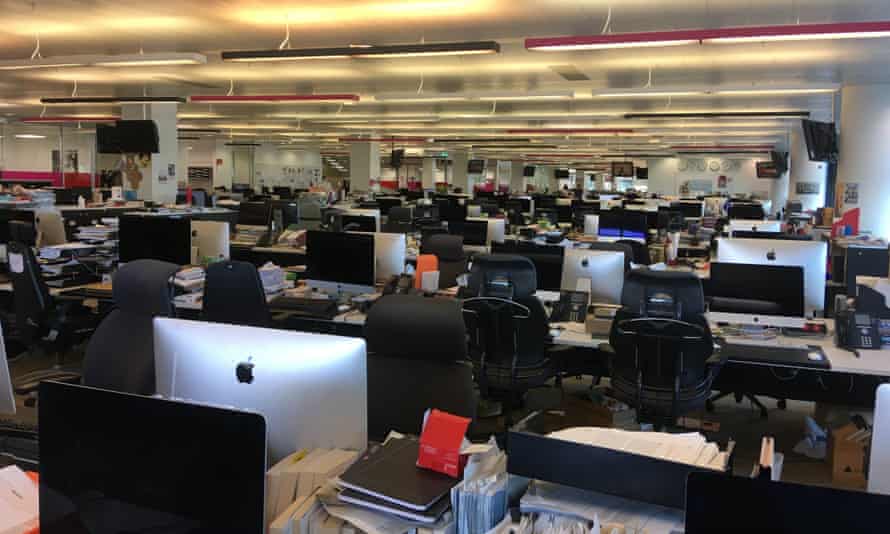 Photograph looking through rows of empty desks with computers under strip lights in The Guardian newsroom