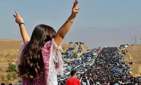 A woman without a hijab, facing away from the camera, holds her arms in the air as she watches protesters march towards Aichi cemetery in Saqez, Mahsa Amini's home