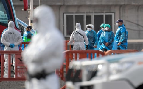 Passengers wearing blue protective suits stand among other people in white protective suits dqxikeidqkikdinv