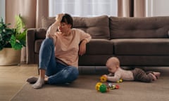 Young tired woman suffering from postnatal depression, holding her head with hand, sitting on floor in living room near her toddler baby playing with toys. New mom tired from motherhood difficulties<br>2M73CKX Young tired woman suffering from postnatal depression, holding her head with hand, sitting on floor in living room near her toddler baby playing with toys. New mom tired from motherhood difficulties