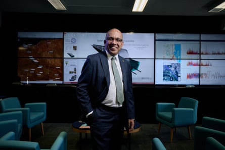 Man in a suit an tie standing smiling in front of a large bank of screens