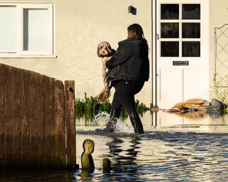 A woman carries a dog in ankle-deep water past a house with the door sandbagged
