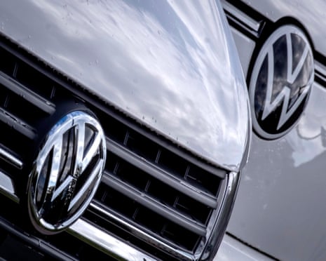 Close-up of the Volkswagen logo on the badge in the centre of the front grille on a silver car, with another logo seen behind on another greyish car to the side