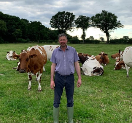 Andrew Hall, who farms in Warwickshire, stands in a green field with a group of cows behind him.