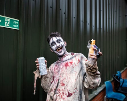 A man in a bloody costume juggles a can of drink, a sausage roll and a vape while smiling maniacally.