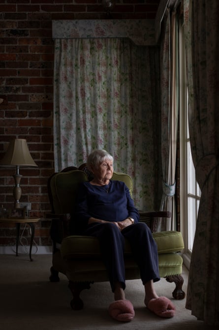 An older woman in a dimly lit room sitting in an upholstered green chair