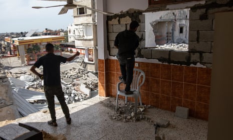 One person looks out at a destroyed city where there was once a wall on a flat, blown out by a missile strike, while another person stands on a chair and rebuilds part of a wall with bricks and cement