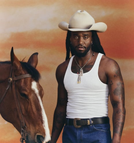 Shaboozey in white vest, blue jeans and cream cowboy hat, posing next to a horse