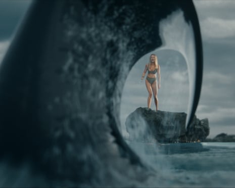 A woman in a bikini stands on a rock as a massive orca whale emerges from the water nearby.
