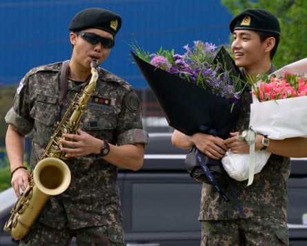 RM plays the saxophone while V looks on at an outdoor sporting facility in Chuncheon in 2025, after their release from military service.