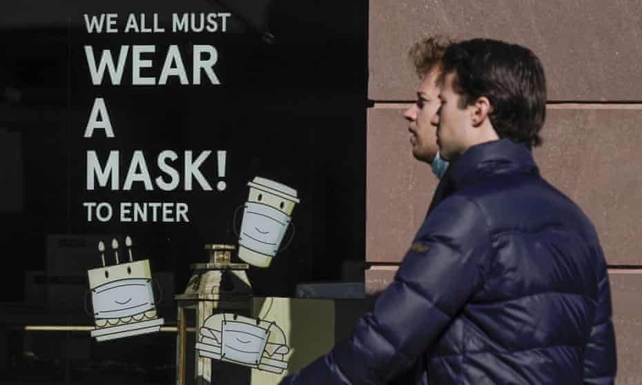 Two unmasked men walk past a sign saying "We all must wear a mask!" with images of a masked cup, hamburger and cake.