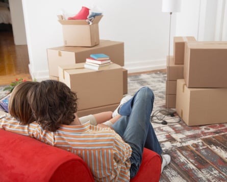 Rear view of young couple on bright red chair in house with cardboard boxes; both have light brown hair; the young man wears a striped cotton shirt and blue jeans but we only see his partner’s hair; there is a bare wooden floor and it looks as if they have just moved into a property together.