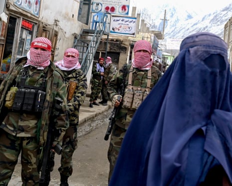 Taliban security personnel with rifles stand guard as a burqa-clad woman walks past
