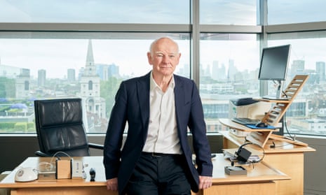 Sir Howard Davies standing leaning against the front of his L-shaped desk with a serious expression and a view of the City of London through the windows behind him
