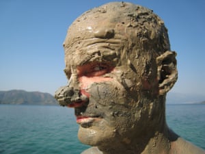 Portrait of man, sea and rocks in background, head and face completely covered in mud.