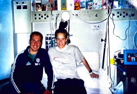 Two men sitting on a hospital bed – one older in blue tracksuit and the other a teen in a white T-shirt, surrounded by lights, wires and get well cards