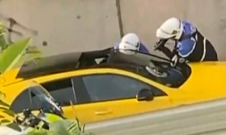 A partial image of a yellow car, with two police officers leaning towards the window on the far side. One officer holds a weapon