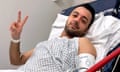 Pouria Zeraati in a hospital gown on an ambulance bed, doing a peace sign