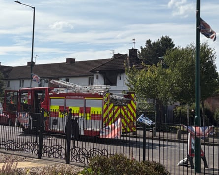 A fire engine is parked in front of a white end of terrace house which has its roof burnt out and fire damage around a window. Union jack flags are flying from lamp-posts and railings in the street.