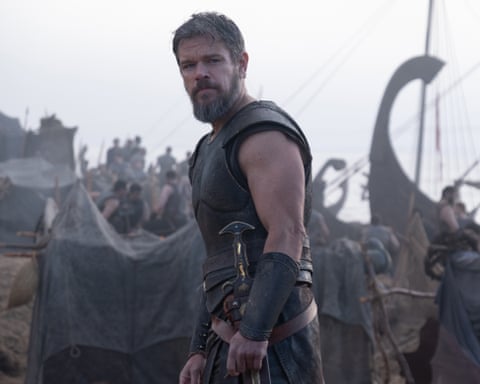 Matt Damon as Odysseus in dark leather armor stands in a misty encampment with Viking ships behind him
