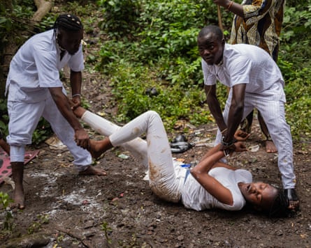 Two young men dressed in white hold on to the arms and legs of a young woman dressed in white who has been rolling around on the ground